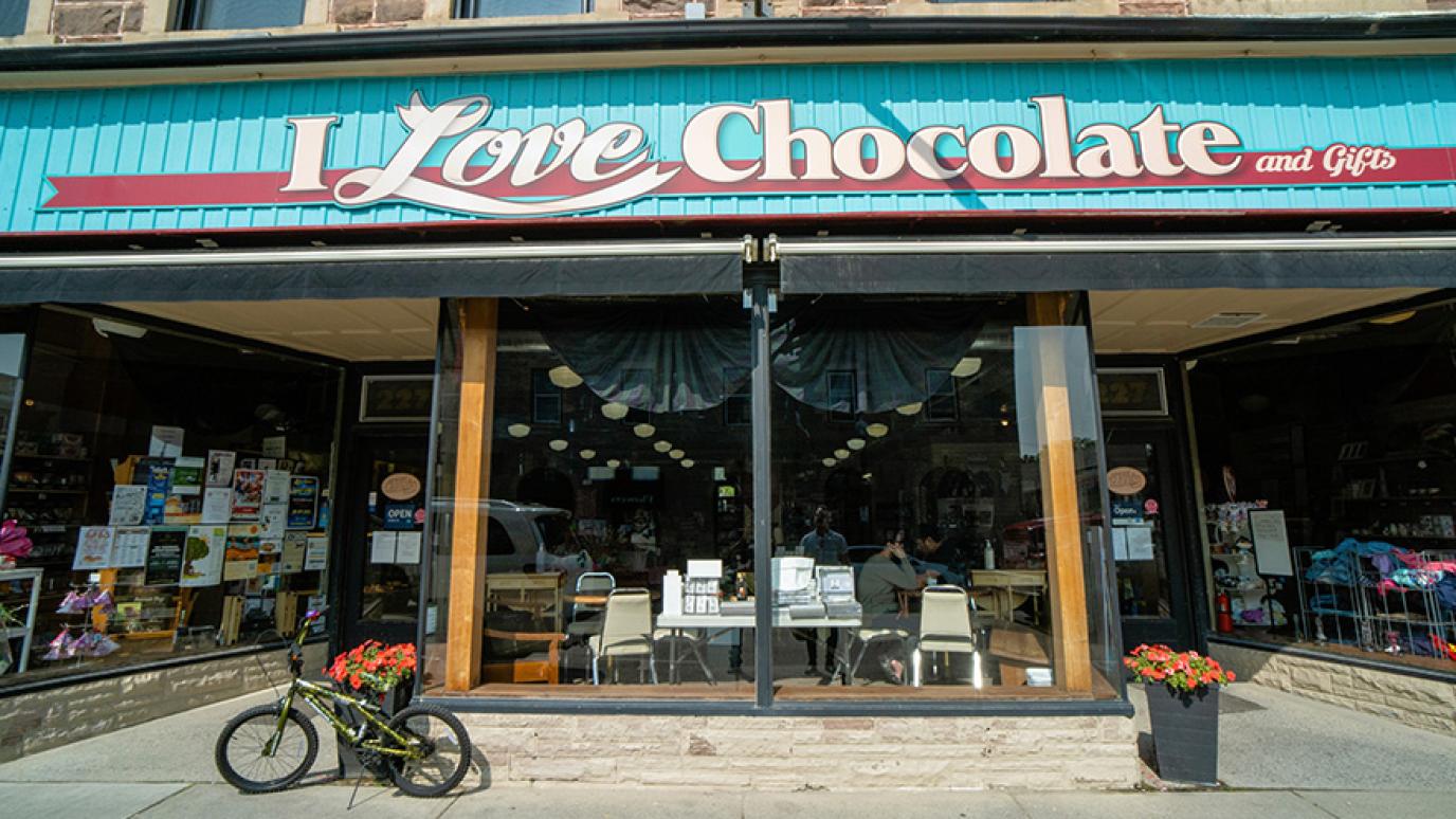 Photograph of a full storefront with a large blue sign with "I Love Chocolate and Gifts" written in white letters. A bike and red flowers sit on the sidewalk out front. 