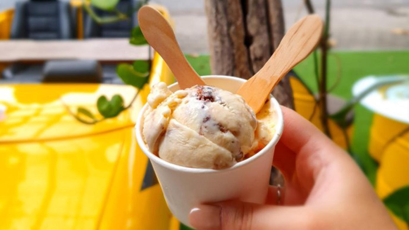 A hand holds up a cup of delicious ice cream with two wooden spoons sticking out of it.
