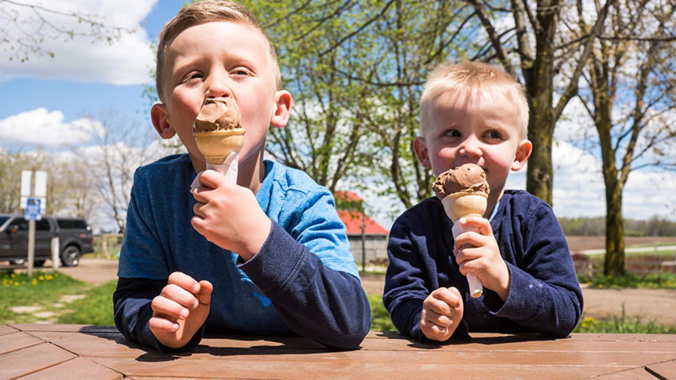 Two blonde haired children in navy blue shirts happily sitting at a table and eating chocolate ice cream cones.