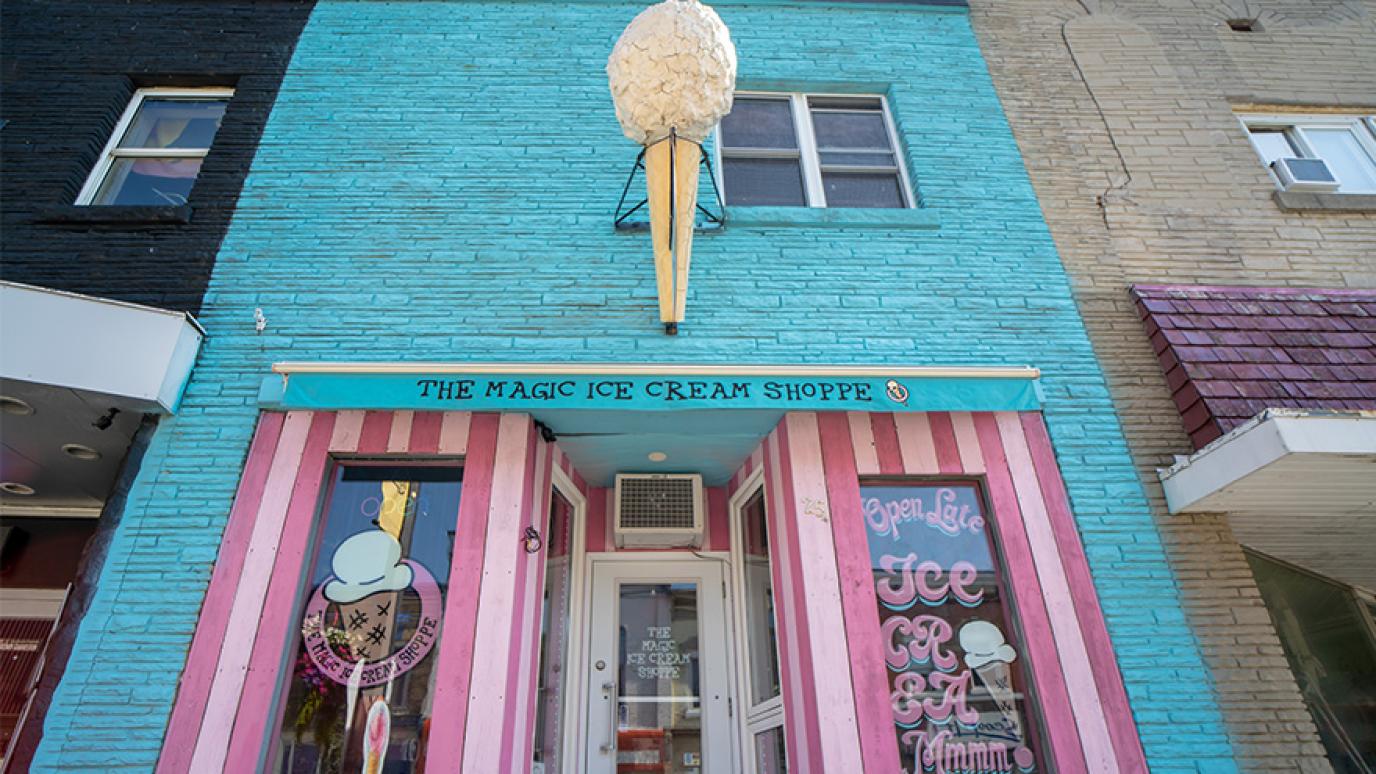 A blue storefront with pink and white stripped window frames, the store name "The Magic Ice Cream Shoppe" and a large ice cream cone sculpture above the door.