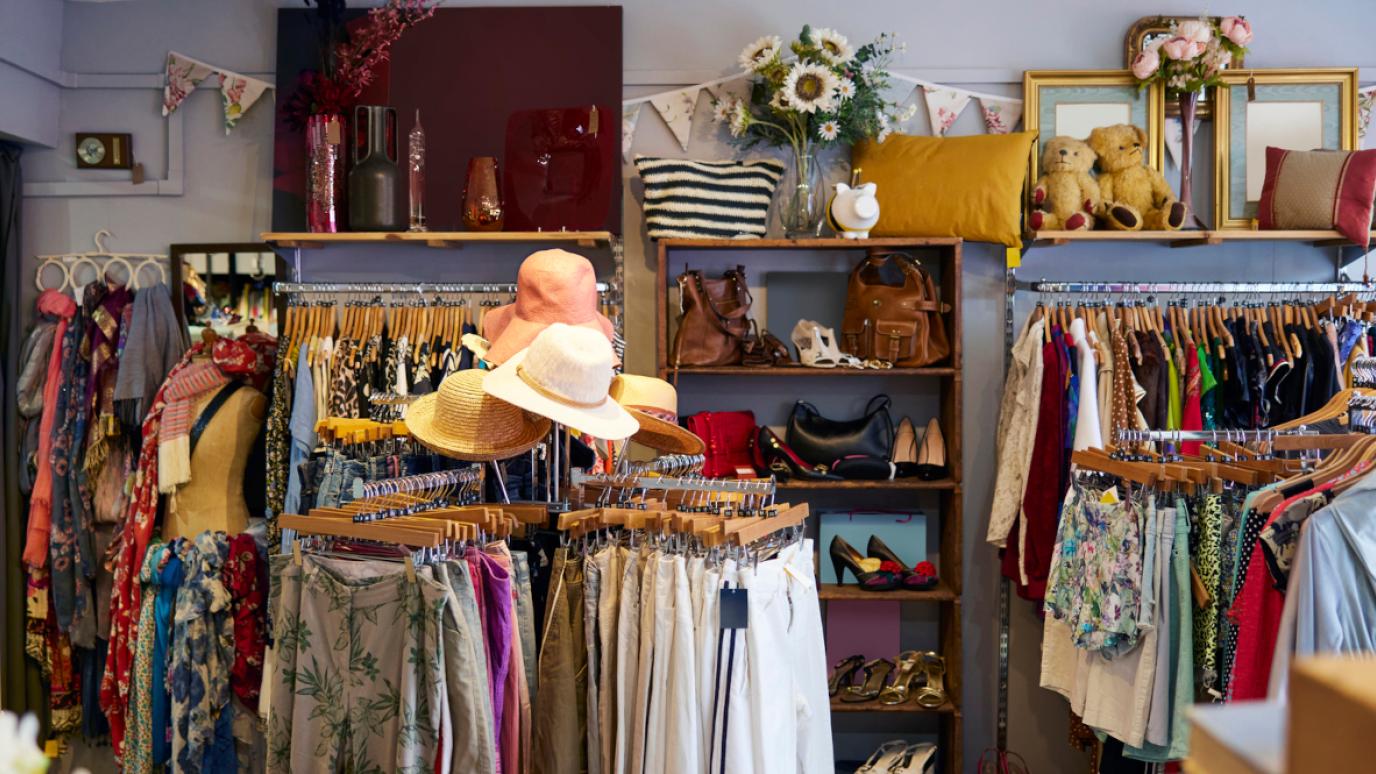 A cozy and colorful thrift store interior with neatly arranged racks of second-hand clothing, hats, handbags, and shoes. The store features decorative items such as pillows, flowers, framed mirrors, and plush teddy bears on upper shelves, creating a welcoming and eclectic atmosphere. A display case on the left showcases jewelry and accessories.