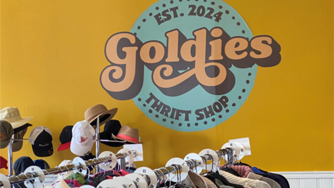The Goldies thrift shop logo displayed on a wall, with a glimpse of nearby aisles featuring clothes, caps and hats.