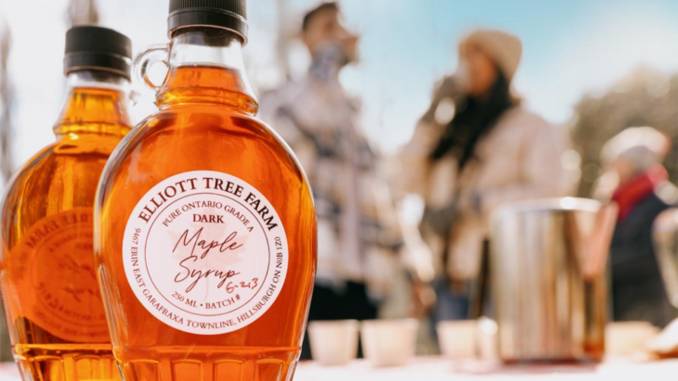 close up of two bottles of maple syrup with people blurred in the background