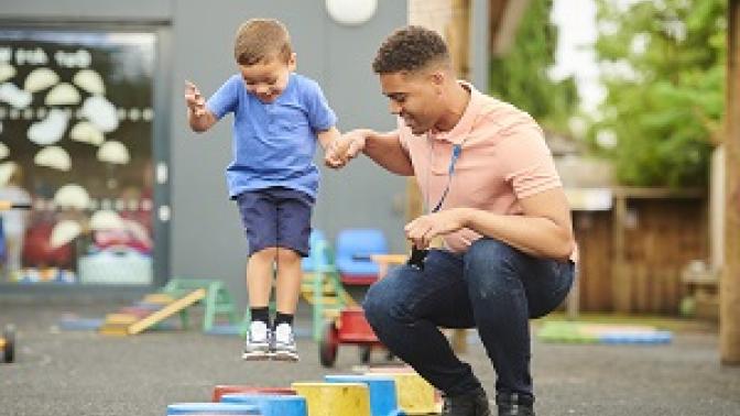 An adult half crouches next to a kid holding their hand as the balance on a beam.