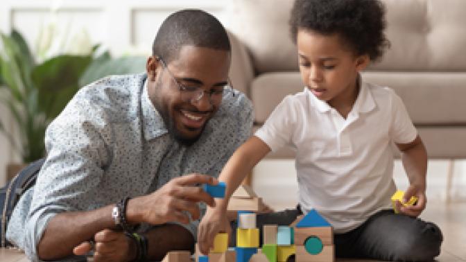 A father lays on the floor next to his son, helping him build a block castle.
