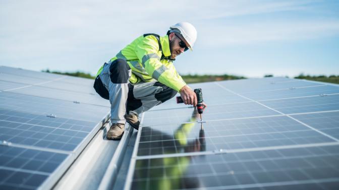 Male worker on a roof using drill to fix solar panel.
