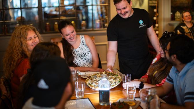 people sitting around table smiling while being served pizza