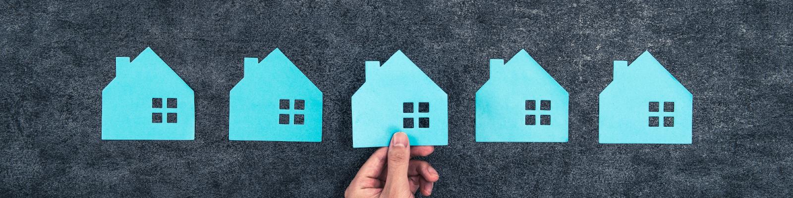 Five simple blue houses cut out of paper. A persons hand is placing the centre one.