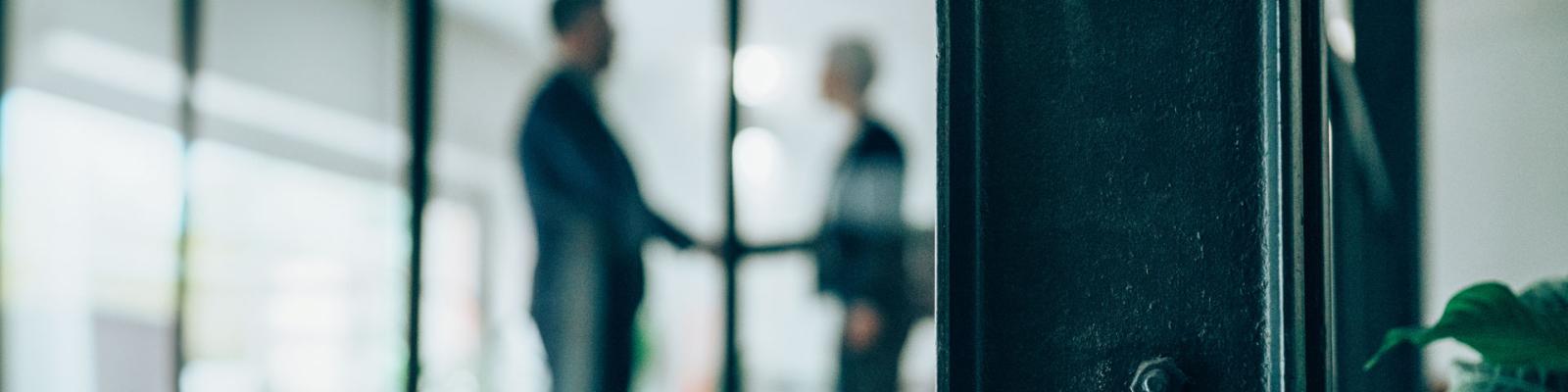 Blurred, cropped image of two office workers shaking hands through closed glass doors