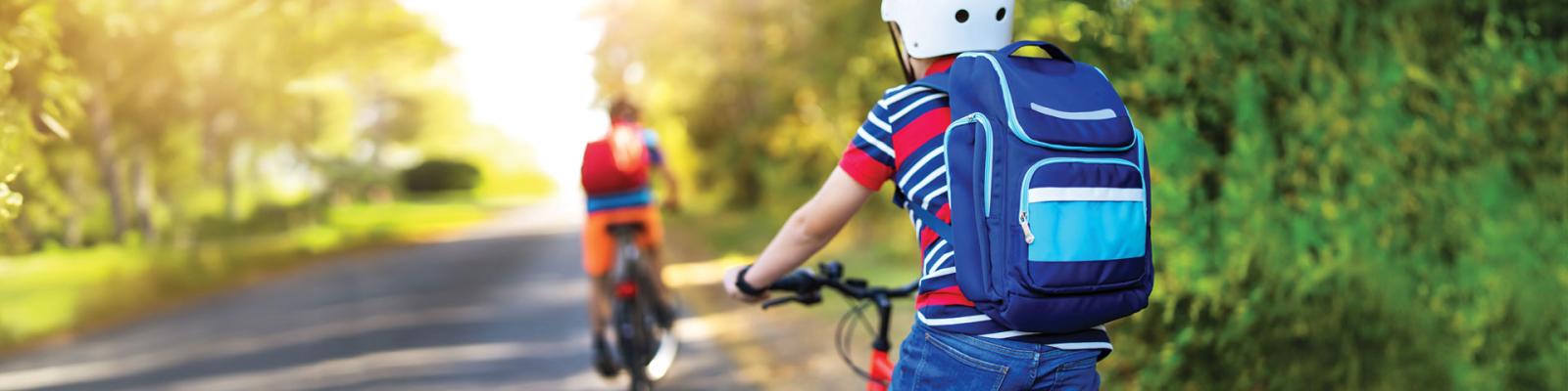 Two kids from behind biking home from school wearing helmets alongside a wooded, rural road