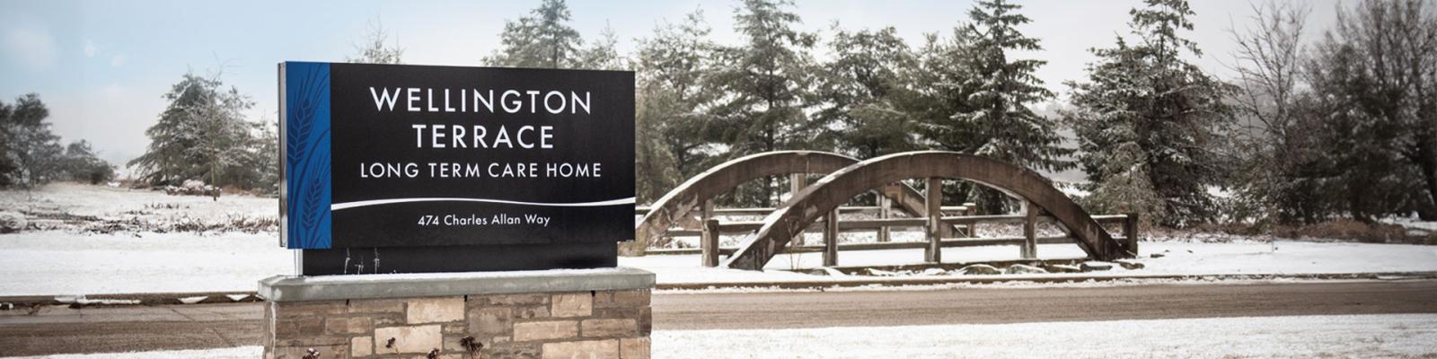 Wellington Terrace Long Term Care Home sign at the front of their entryway, a stone bridge and forest area in the background
