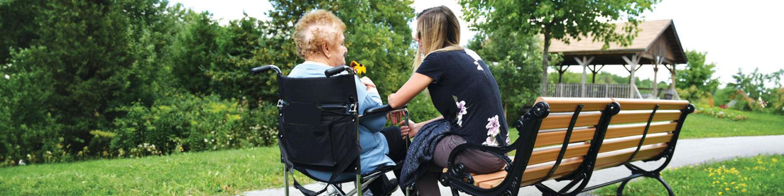 Elderly woman in wheelchair visiting with young woman on a bench outside enjoying a sunny day