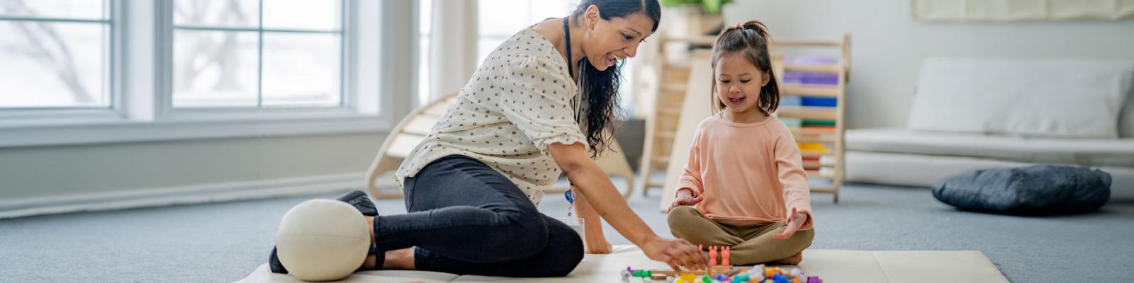 A child care worker sits on the floor with a little girl playing together with colourful figurines