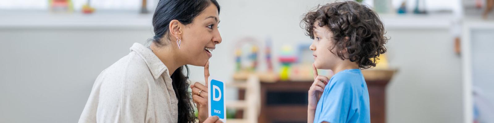 A female speech therapist works with a young boy as they practice their sounds and letter enunciation. The therapist is holding up a cue card with the letter D and the word "Duck".