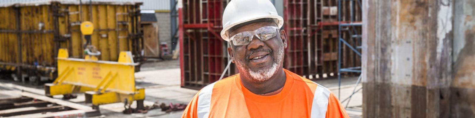 Close up of a older construction worker on a worksite wearing safety glasses, a white hard hat and orange safety vest