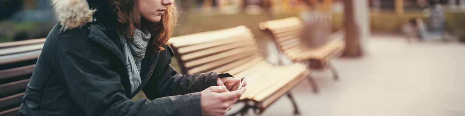 Teen girl looking concerned sits on a park bench in a winter coat looking down at her cell phone