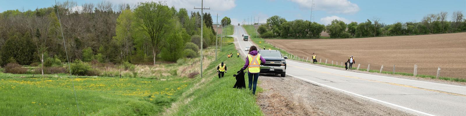 A group of people wearing reflective vests pick up garbage from the sides of a County road.