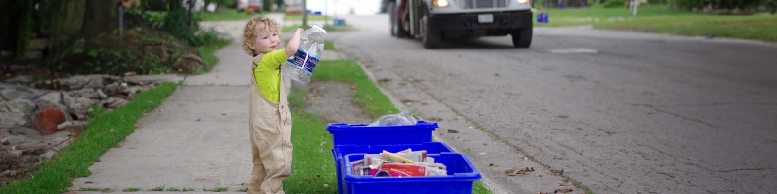 Boy adding a plastic bottle to his blue box as the garbage truck arrives at the curb