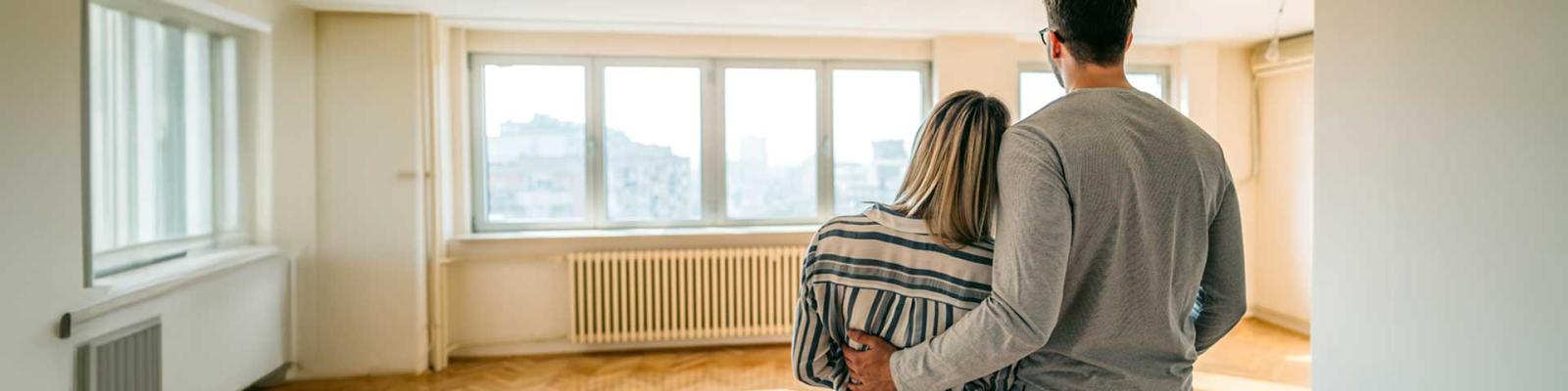 The back of a man and woman standing together hugging while looking at their empty, bright apartment. 