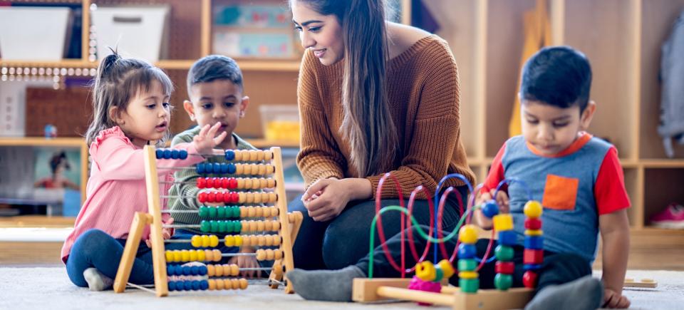 A female teacher of Middle Eastern decent sits on the floor with a small group of students as they learn to count together with an abacus. They are each dressed casually and have neutral expressions on their faces as they work together.