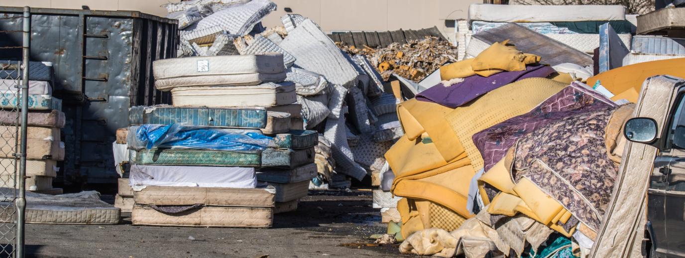 A pile of mattresses both whole and deconstructed piled beside a large waste bin outside at a recycling facility. 