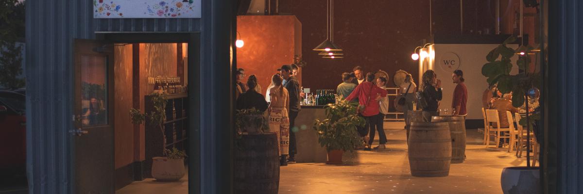 A warmly lit indoor space with people gathered around tables and barrels in a cidery, seen through a large open doorway.