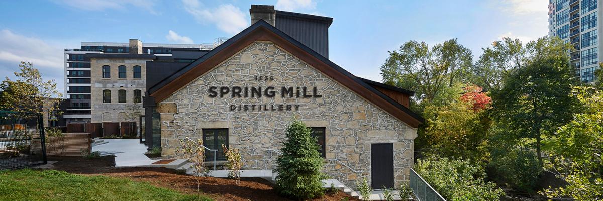 A stone building with "SPRING MILL DISTILLERY" written on its facade stands with trees and a modern building behind it.